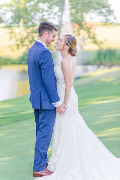 A groom and bride hold hands and look at each other while standing next to a pond on a golf course.