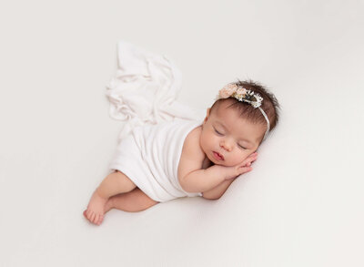 Baby girl sleeping atop of blush tulle is wrapped in a matching blush wrap with her arms and legs exposed. She is curled up laying on her back and facing the camera. Captured by top NYC Newborn Photographer Rochel Konik Photography.