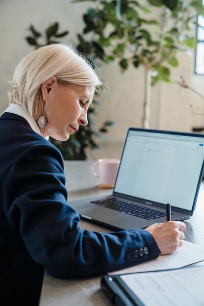 An older white woman working at a laptop and writing in a notebook