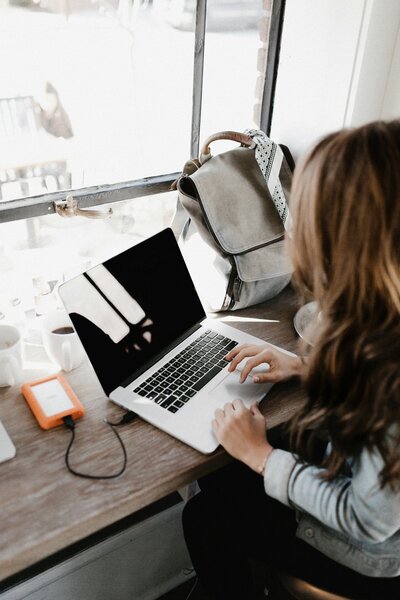 The productivity in motion as a blond woman works diligently on her grey laptop. The setup includes a beige backpack beside her, highlighting mobility and efficiency. The laptop is seamlessly connected to a sizable USB, showcasing the expert's commitment