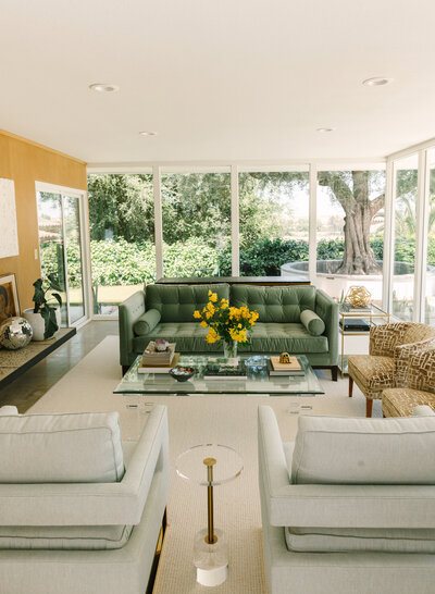 Bright mid-century modern living room featuring a green velvet sofa, glass coffee table, modern accent chairs, and floor-to-ceiling windows with lush greenery outside — photographed by Dani Lacey in Greenville SC.