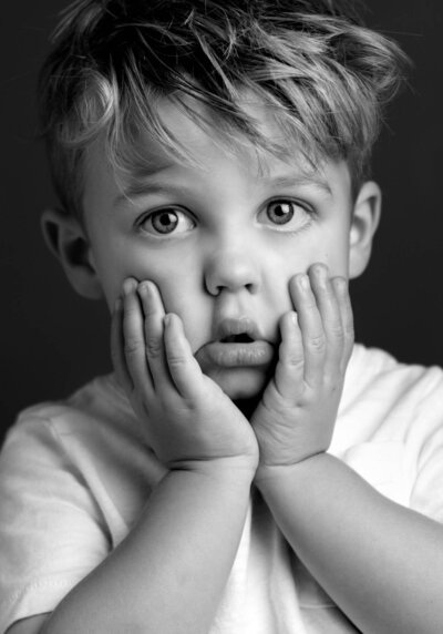 A young boy holds his hands to his face, surprised, with a playful expression

