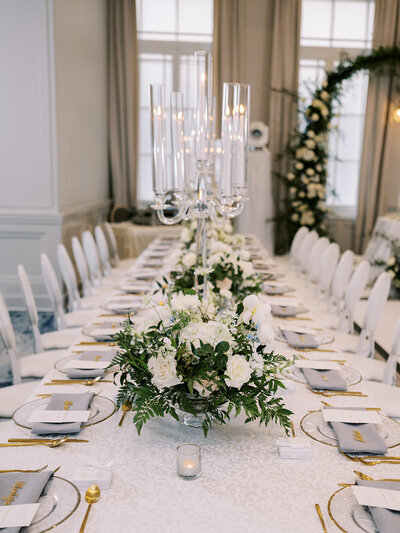 A long, elegantly set dining table with white tablecloths, white floral centerpieces, gray napkins, and gold cutlery orchestrated by a top Calgary wedding planning team. A tall candelabra centerpiece and an arch decorated with flowers grace the background.