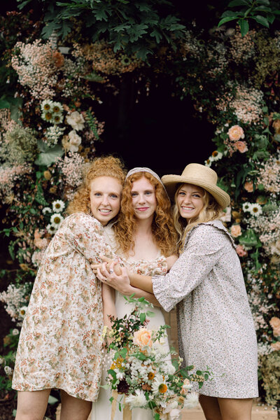 Bride Peyton Curry wears an elegant Doen gown while embracing her sisters, who are dressed in floral Doen bridesmaid dresses, during her intimate backyard wedding in Richmond, Virginia. A candid moment captured in film-style wedding photography by a documentary-style wedding photographer.