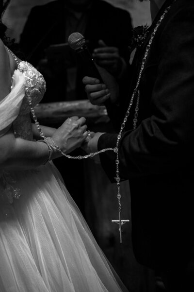 A close-up of a bride and groom holding hands, entwined with a rosary, during a wedding ceremony, exuding love and commitment.