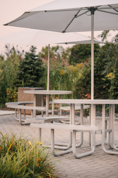 Two white tables with benches and umbrellas on a brick patio, surrounded by lush landscaping