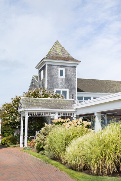 Rhode Island Wedding Photographer | Kelsey Sheehan Photography Timeless Rhode Island Weddings | Charming coastal house with a shingled tower and lush landscaping, featuring ornamental grasses and flowering shrubs under a partly cloudy sky.