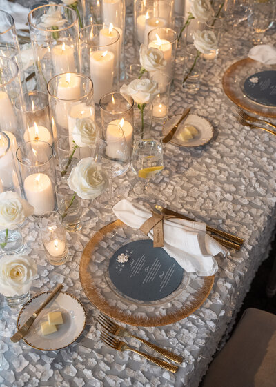 Textured floral linens, with gold lined flatware and dozens o white candles and white roses