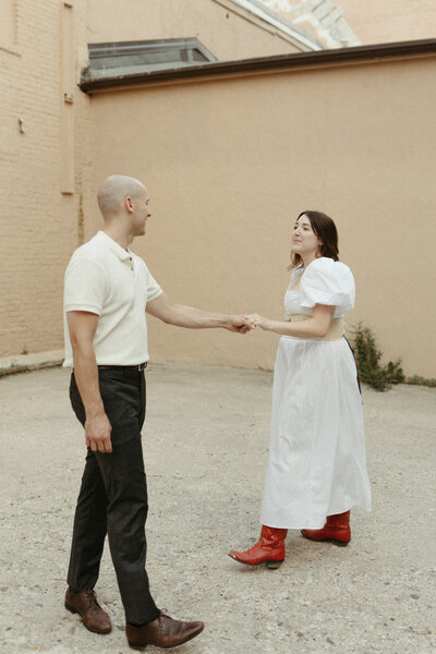 A stylish couple holds hands during an engagement session, featuring the bride-to-be in red cowboy boots.
