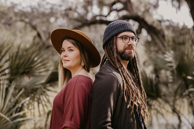 Man with beanie and dreads stands back to back with his wife as she also looks opposite of him while wearing a wide brimmed hat. They look happy, yet ready to take on new adventures.