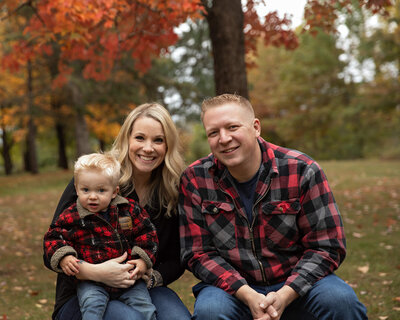 Couple with their toddler boy sitting on a bench with beautiful fall trees in the background.