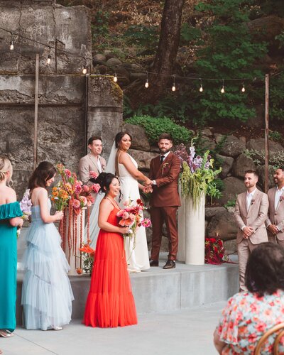 A candid wedding photo during the ceremony. The couple is standing on a stage along with their officiant. They are holding hands. Their wedding party, dressed in colorful attire, is standing a step down from the couple. There are bright color wedding flowers on either side of the couple on cream pedestals. The setting is The Ruins in Hood River.