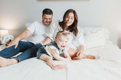 a father helps his toddler daughter stand on the bed in their brightly lit bedroom