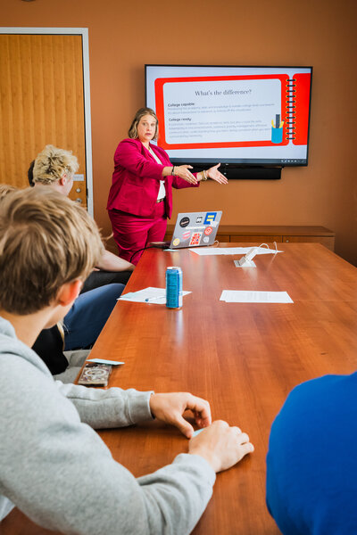 Mya Andersen, College Coach & Student Success Expert, presents to a small group in a meeting room with a slide on a screen behind her. She leads the discussion wearing a red suit, sharing insights and strategies for academic achievement.