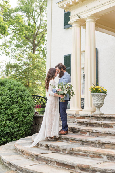 A bride and groom huddle close on the steps of a manor house