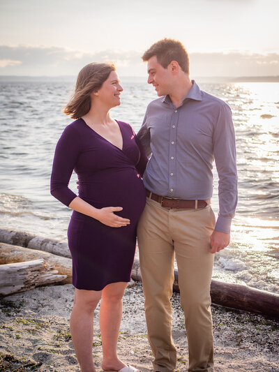A couple standing along the shoreline  at Carkeek Park in Seattle look fondly at each other  as the pregnant woman hold her protruding belly.
