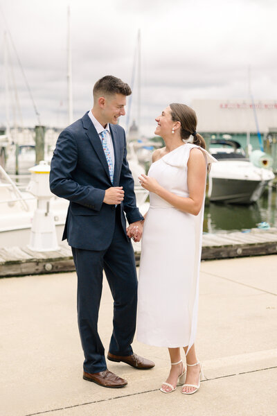 A couple stands on a dock by the water, holding hands and smiling at each other. The man is in a navy suit with a patterned tie, and the woman is wearing a white dress. Boats and a cloudy sky are in the background.