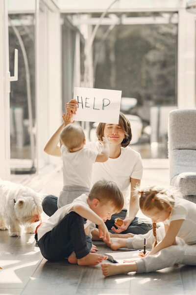 A tired mom playing with her three children on the floor, holding a sign that reads "help". 