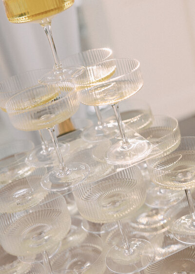 Champagne tower being poured during a wedding reception, featuring stacked coupe glasses captured in soft, golden light.