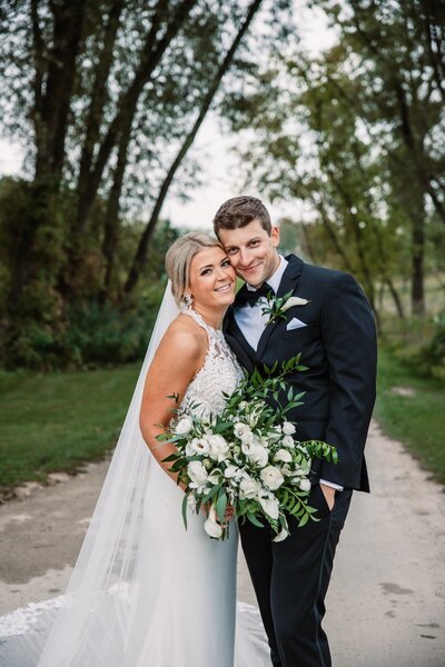 bride and groom smile cheek to cheek at the camera while holding a large flower bouquette of while and greens