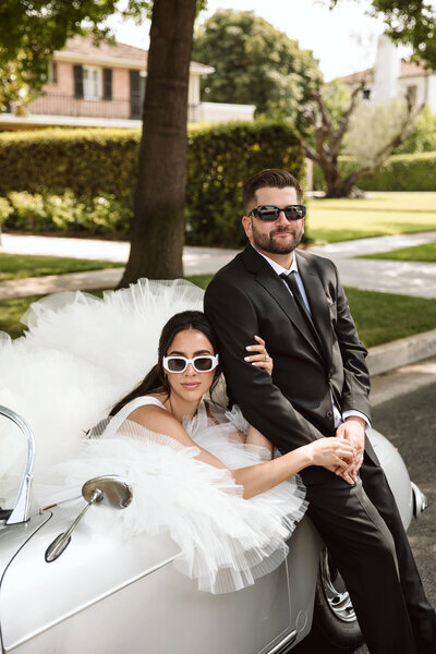 Kohnur Bride in modern glam makeup with voluminous lashes and nude lips wearing a tulle gown and white sunglasses beside the groom in a black suit, sitting on a vintage car in Los Angeles.