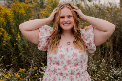 Girl smiles with both hands in her hair wearing a floral dress in the middle of a field.