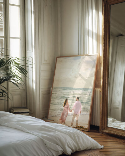 Framed maternity photograph of a couple holding hands on the beach, styled in a sunlit Parisian bedroom with classic panel walls and a gilded mirror.