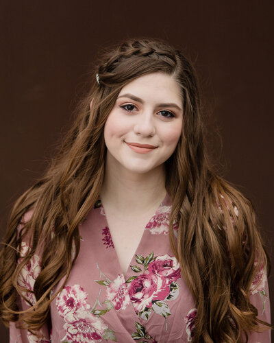 Senior teen girl with long brown hair and braid in floral dress against brown wall