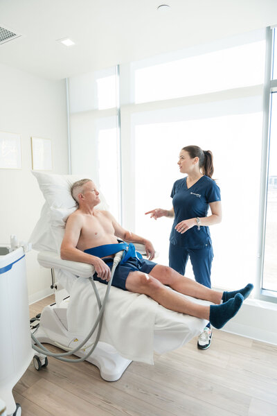 est CoolSculpting Dallas services with an expert performing the treatment. The professional, dressed in a blue brand uniform, is working with a male patient lying on a white stretcher next to a CoolSculpting machine.