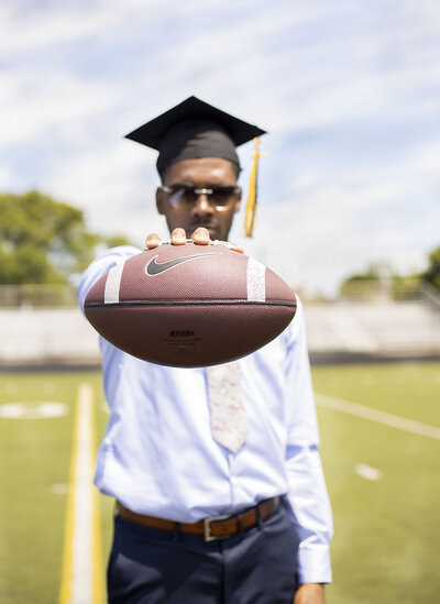 Senior boy with light blue dress shirt, navy blue slacks, and sunglasses wearing black graduation cap holding a football on the football field