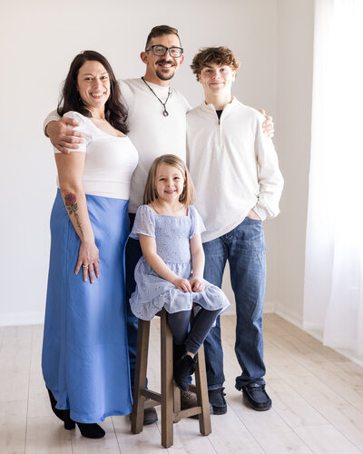 A family of four.  Mom, Dad, and high school aged brother standing, with young sister sitting on a stool. Mom is wearing a blue skirt, white shirt with dark brown hair.  Dad is wearing a white long sleeve shirt and blue jeans, with dark brown hair.  Son is wearing a light tan shirt, blue jeans shorts with brown hair.  Daughter wearing a purple shirt with blue jean leggings.