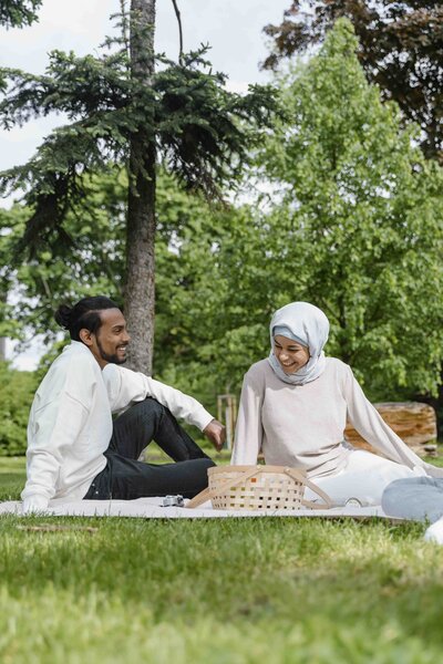 Two people sit on a picnic blanket in the grass, with trees on the background. They smile toward each other, over the picnic basket between them. The person on the left is masculine presenting, with dark skin and long black hair pulled into a bun, and the person on the right is feminine presenting, wearing a light colored hijab and long-sleeved shirt.
