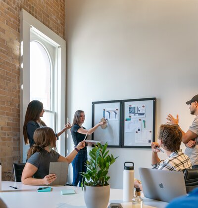 Three people are gathered around a laptop in an office setting, looking at the screen. One person is seated and appears to be explaining something, while the other two are standing and listening attentively, embodying educational leadership in this post-secondary environment.