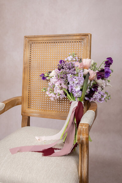 Purple, pink, and white bridal bouquet wrapped with a rose colored ribbon sitting on a linen and wicker chair