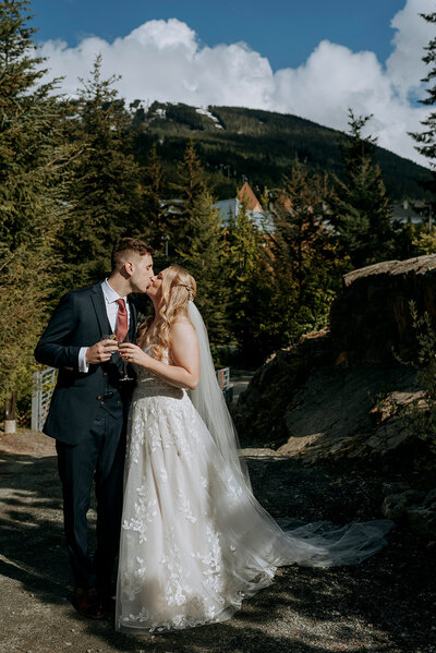 A bride and groom wearing a yellow and green puffy jacket hold each other on top of Rainbow Glacier in Whistler. There is snow all around and blue skies.