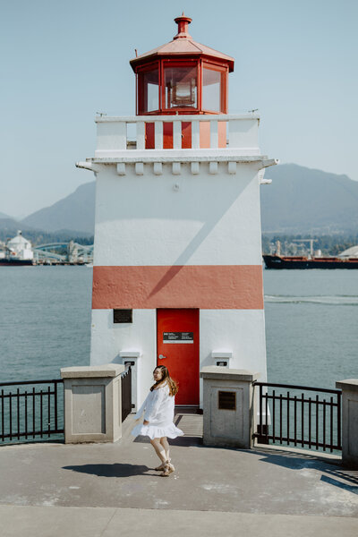Anne, an Asian Woman, is at the Stanley Park Lighthouse in Vancouver, BC, Canada smiling and twirling as the wind lifts the ruffle of her white knee length dress. She is wearing suede espadrilles.