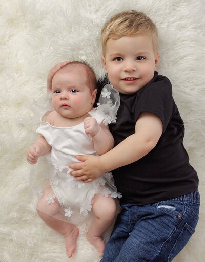 Big brother cuddling his newborn baby sister on a soft white blanket during an in-studio lifestyle newborn session in Bergen County, NJ