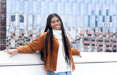 Senior girl wearing dark tan corduroy jacket, white cowl neck sweater, and stonewash destructed jeans on rooftop