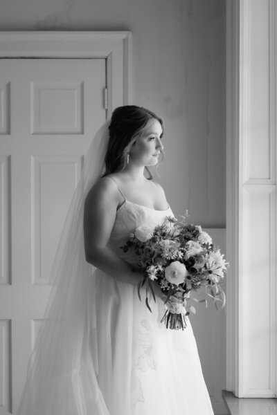 black and white portrait of a bride holding her bouquet of flowers looking out of a window