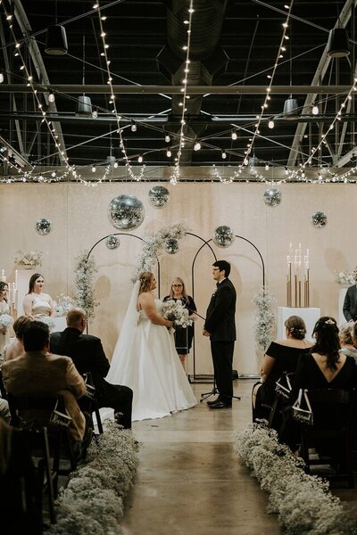 Wichita bride and groom smile at each other at the alter during their ceremony with overhead décor of disco balls and string lights