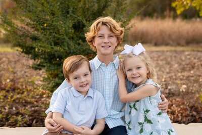 Three smiling children sitting close together outside, dressed in light colors, surrounded by trees and fall foliage, part of the Munsey family.
