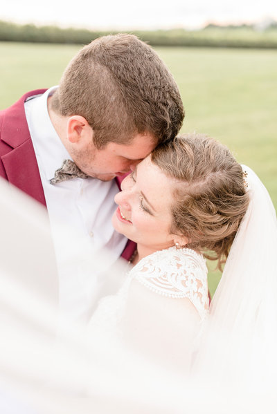 Groom hugging his Bride tightly as his face rests on her right cheek with her long veil blowing in front of them.