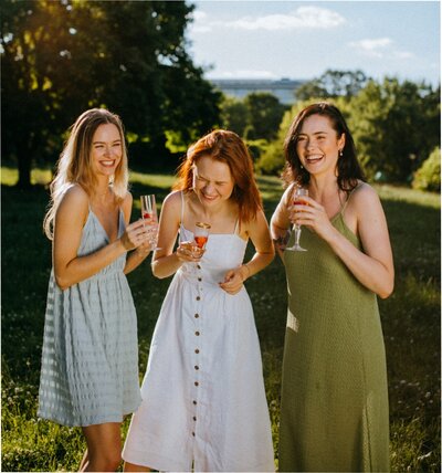 A group of young women with champagne flute glasses in their hand laughing together with nature in the background