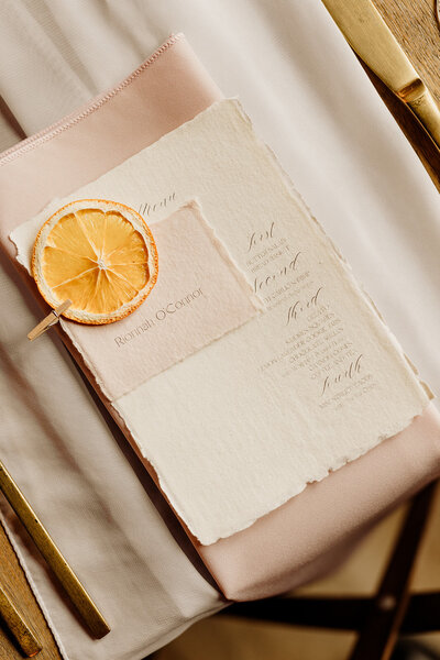 Invitation suite flatlay with textured canvas paper and ripped edges and elegant script writing. Dried orange lays atop.