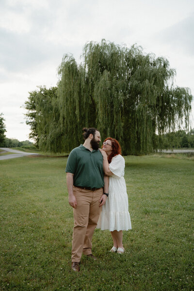 photo of engaged couple hugging and looking into each others eyes lovingly with a weeping willow tree in the background