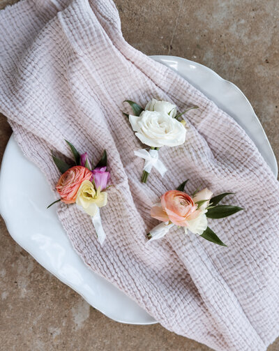 Flat-lay photograph of three colorful spring wedding boutonnieres arranged on a soft textured blush cloth over a white plate, featuring pastel ranunculus, roses, and lisianthus with greenery—styled wedding detail image showcasing romantic floral design for modern celebrations.