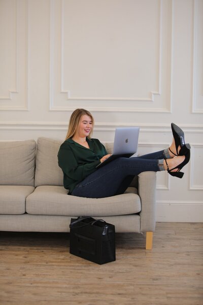 Woman wearing crossing legs and laying back on a gray couch with black heels hanging over the arm of the couch and an open silver laptop on top of her legs.