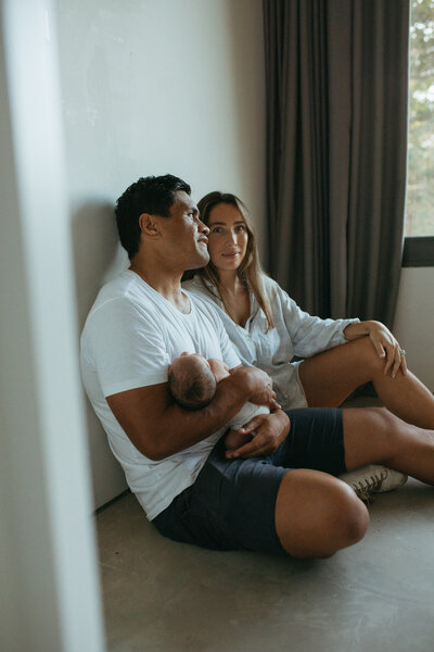 In home newborn Photoshoot , a mother and father sita holding their newborn and the mother is glancing at the camera through a doorway- Photo by Rebecca Rottcher| Newborn Photographer
