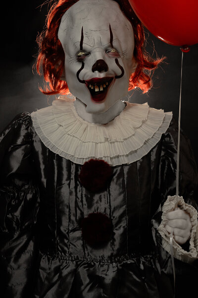 Person wearing Pennywise the Clown costume holding red balloon. Illuminated from behind to look scary.