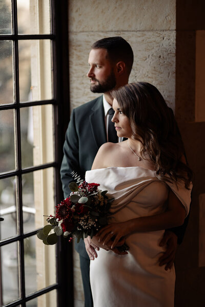 Bride and groom posed for a portrait near a indoor window. Groom is kissing the brides temple.